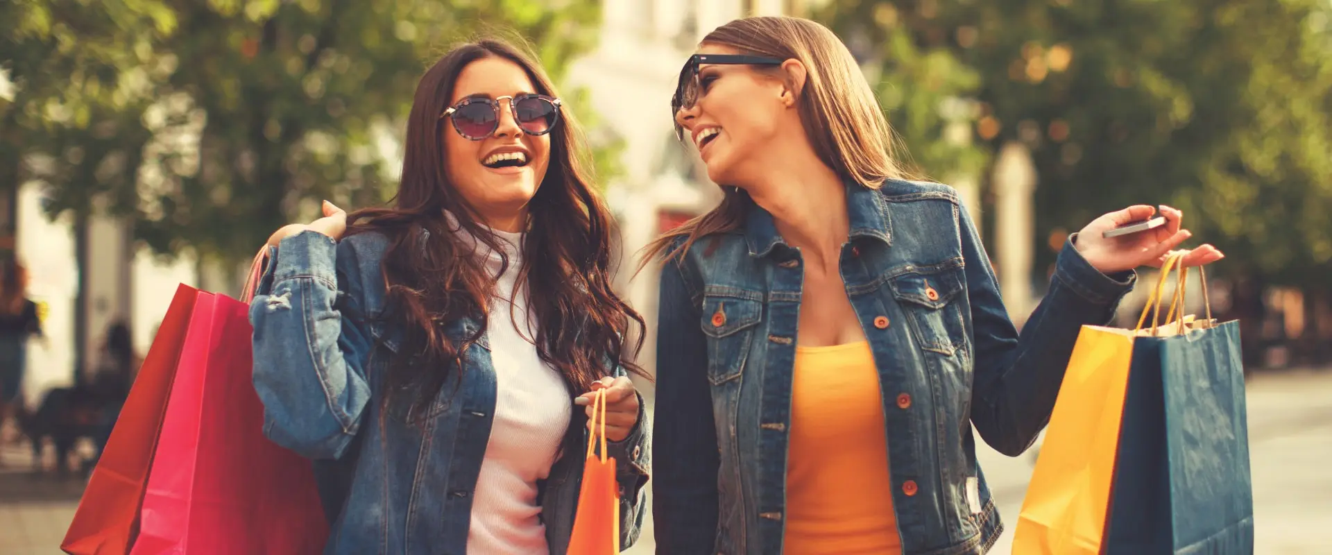 Sand-Creek-Website-Banner-2025-Jan-min Two woman wearing denim jackets, glasses, and holding shopping bags are laughing together in the sunlight.